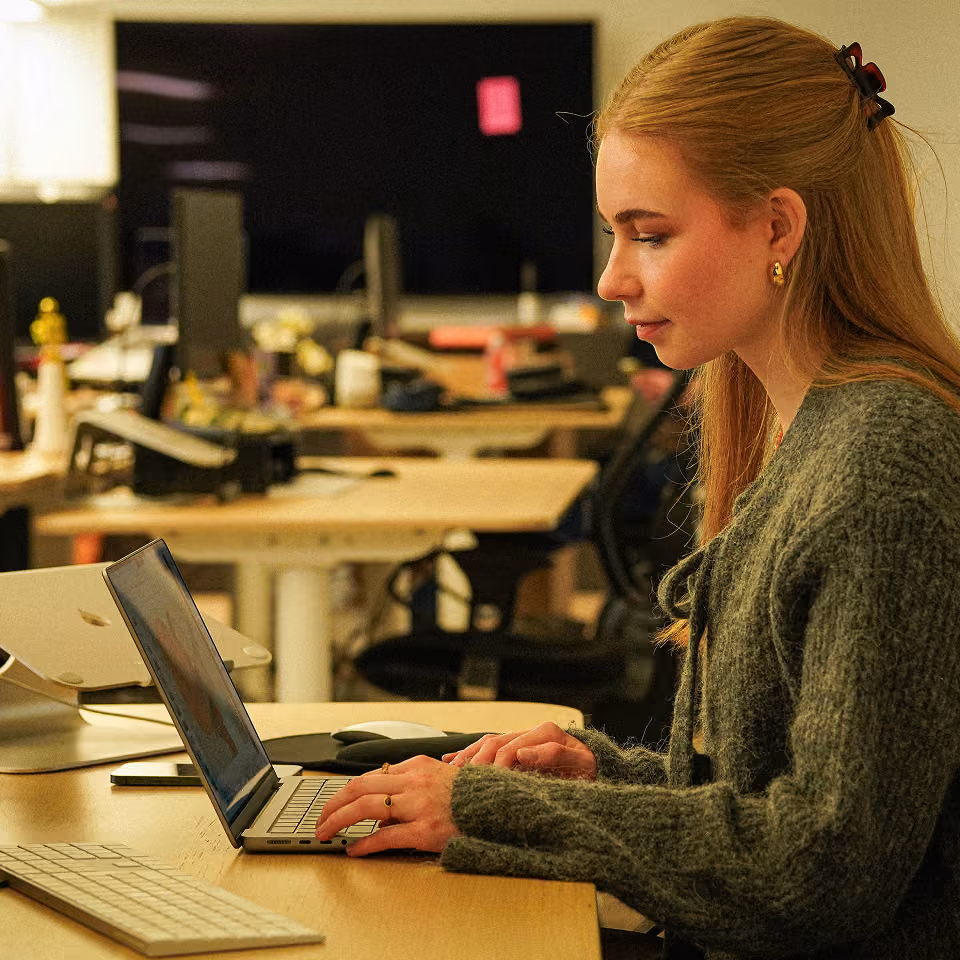 Junge Frau mit langen blonden Haaren, die an einem Laptop in einem modernen, hell beleuchteten Büro arbeitet.