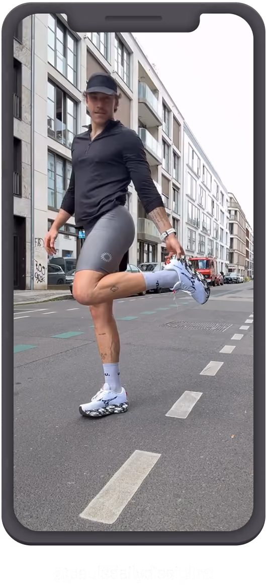 Ein sportlicher Mann in schwarzem Langarmshirt, grauer Sporthose und weißen Laufschuhen dehnt sich auf einer Stadtstraße, indem er einen Fuß hinter sich greift. Er trägt ein schwarzes Käppi und steht auf einem abgetrennten Fahrradweg vor modernen Wohngebäuden.