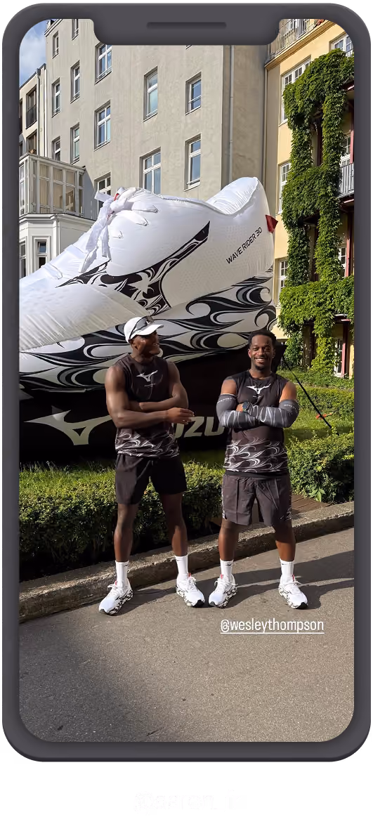 Zwei Männer in Sportkleidung stehen vor einem großen aufblasbaren weißen Sportschuh mit schwarzem Muster und der Aufschrift 'Wave Rider 30', umgeben von Gebäuden und grünen Pflanzen.