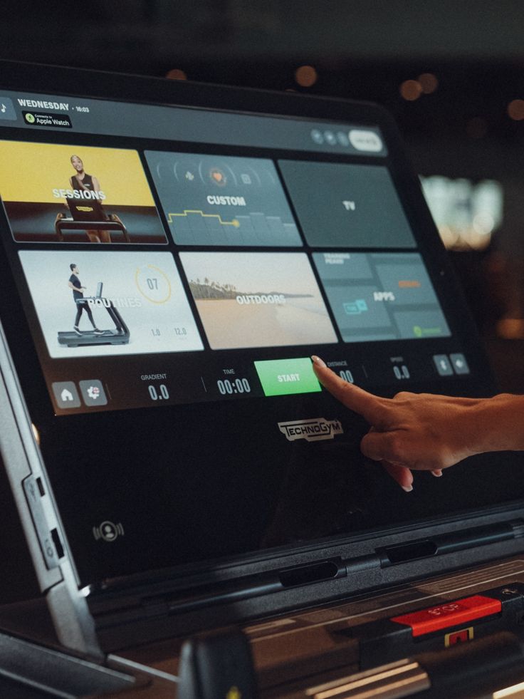 A person’s finger pressing the green START button on a Technogym treadmill touchscreen displaying workout options.