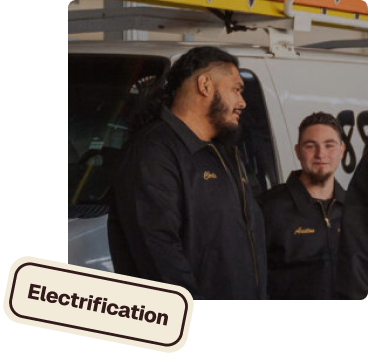 Two men in black work uniforms standing in front of a white van, one looking at the other and both smiling, with a tilted sign labeled Electrification in the foreground.