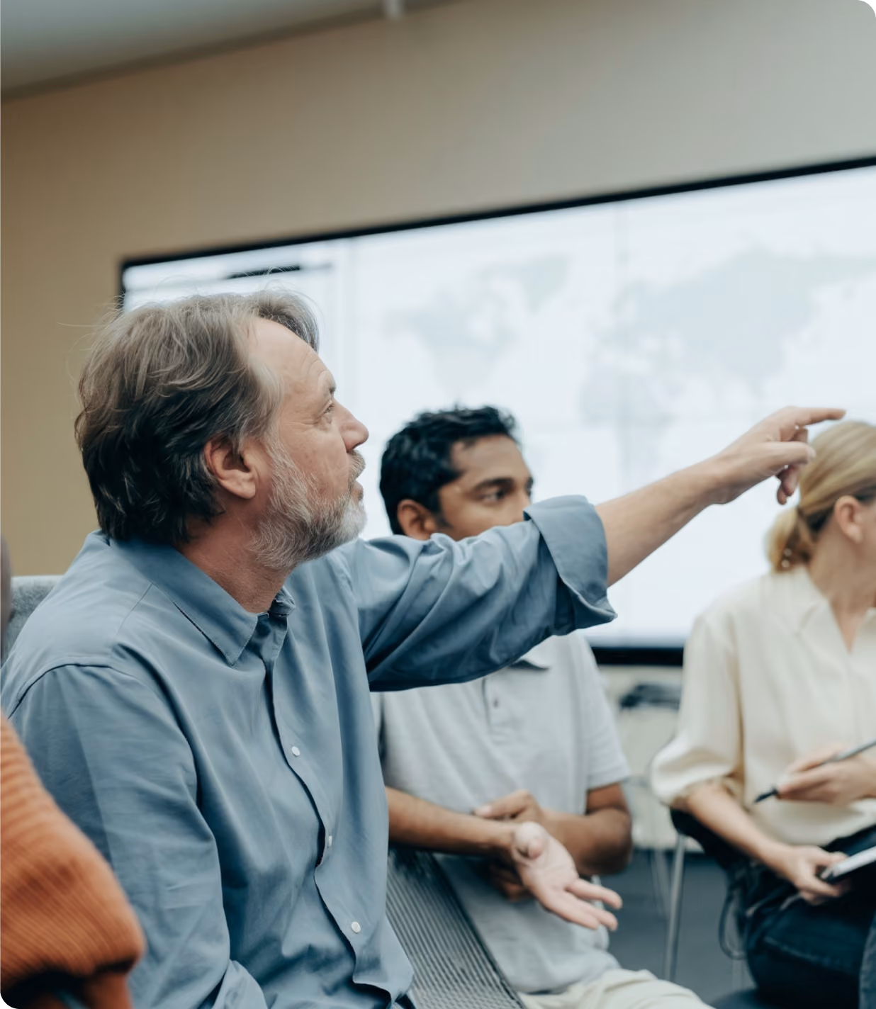 Man with gray hair and beard in a blue shirt pointing while sitting in a meeting room with two other people and a large screen displaying a world map in the background.