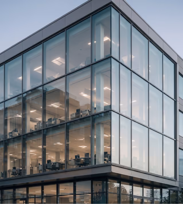 Modern office building with large glass windows showing empty workstations inside during early evening.