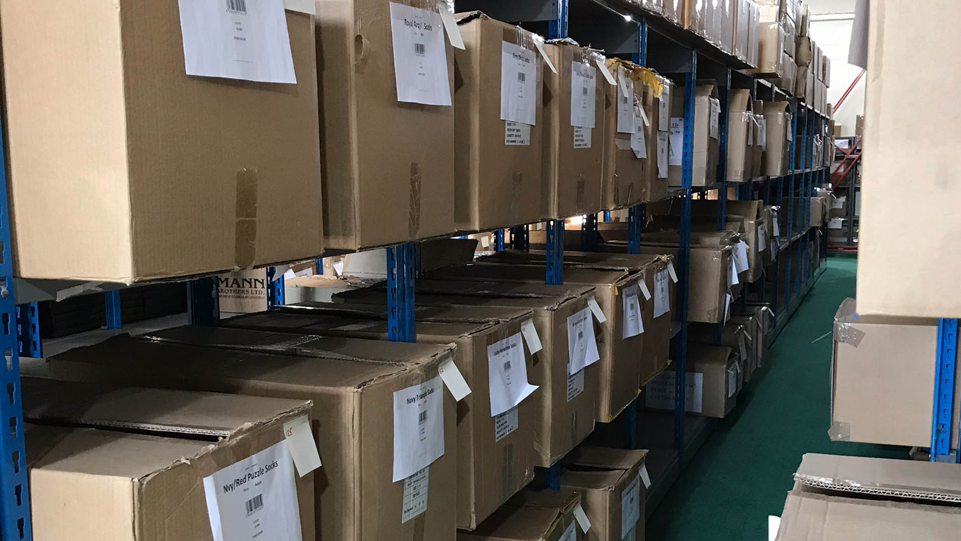 Labelled merchandise cartons stored on warehouse shelving beside a stockroom aisle.