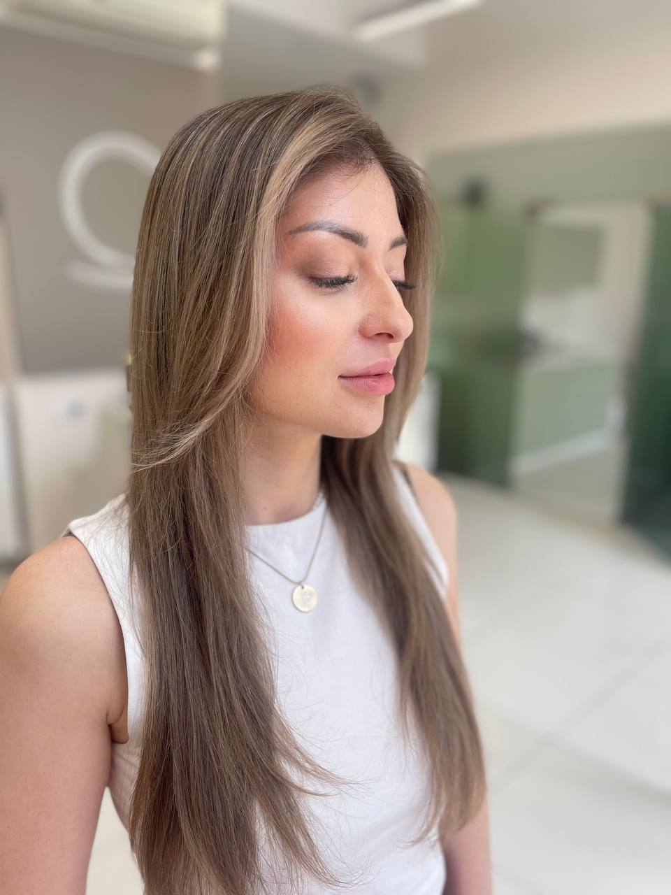 Side profile of a woman with long, straight, light brown hair wearing a sleeveless white top and a round pendant necklace, standing indoors with a softly blurred background.