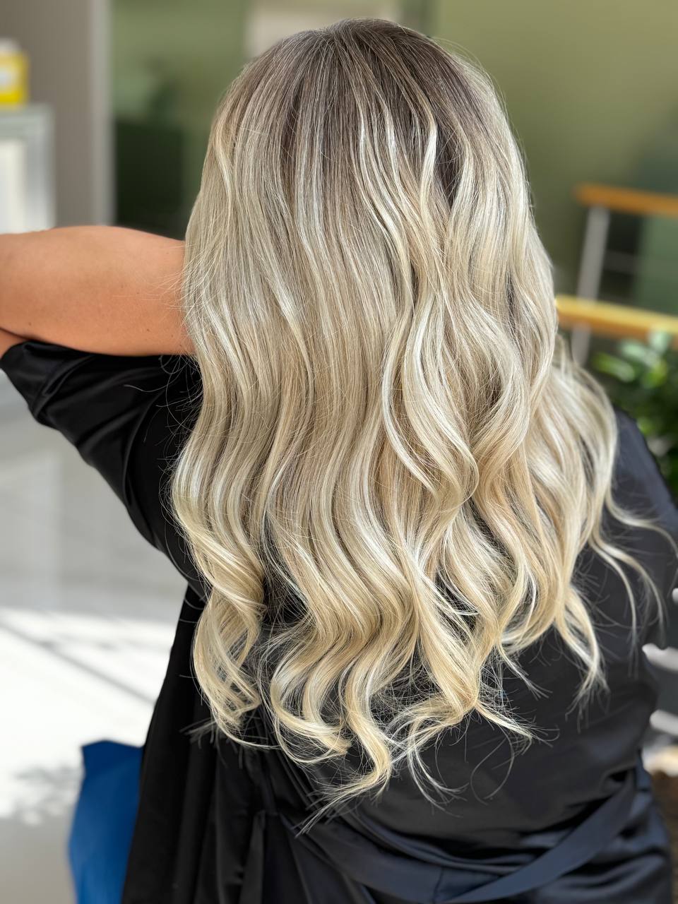 Back view of a person with long, wavy, blonde hair wearing a black top in an indoor setting.