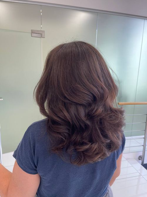Person viewed from behind with medium-length, wavy brown hair, wearing a dark blue shirt in an indoor setting with glass panels and metal railings.