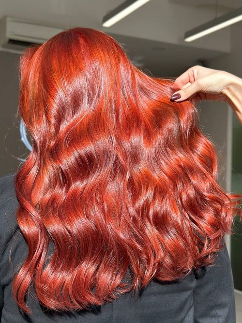 Back view of long, shiny, wavy red hair held slightly by a hand with dark polished nails, against a blurred indoor background.