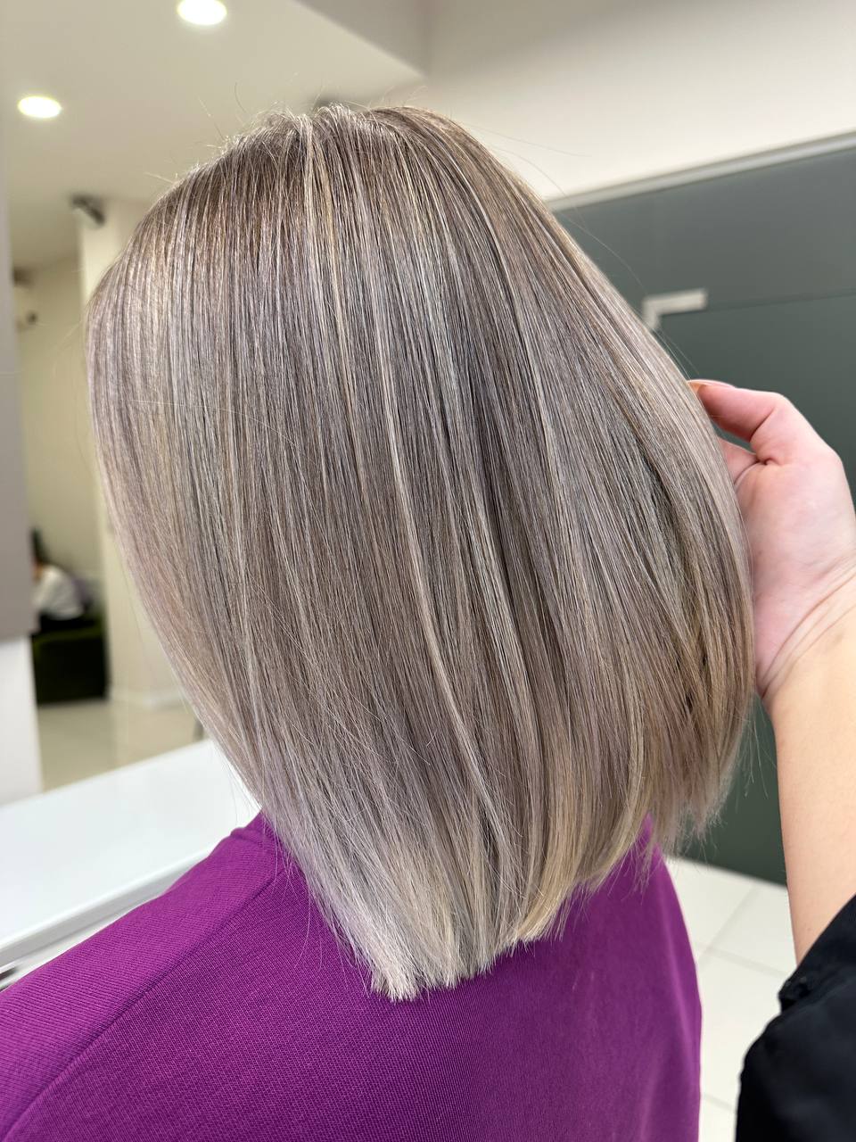 Close-up of a person with smooth, straight, shoulder-length ash blonde hair with subtle highlights, wearing a purple top, with a hand gently holding the hair on one side.