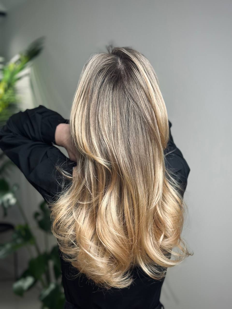 Person with long, voluminous, wavy blonde hair shown from the back, wearing a black top against a neutral background.