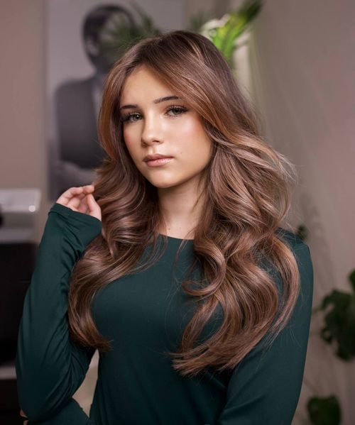 Young woman with long, wavy light brown hair wearing a dark green top, posing indoors with a neutral expression and holding a strand of hair.