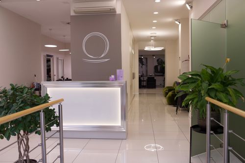 Modern reception area with illuminated white front desk, gray feature wall with circular logo, white tiled floor, green plants on either side, and green chairs along a hallway leading to rooms in the background.