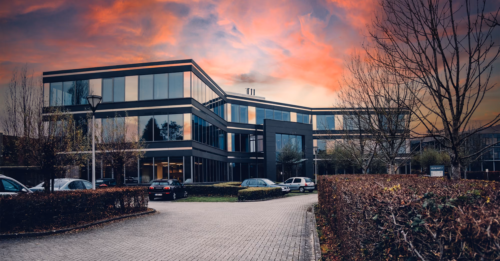 Modern office building with large glass windows and black framing under a colorful sunset sky, surrounded by leafless trees and parked cars along a paved driveway.