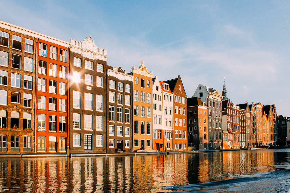 Row of colorful, narrow canal houses along a waterway in Amsterdam under a clear sky, with reflections in the water.