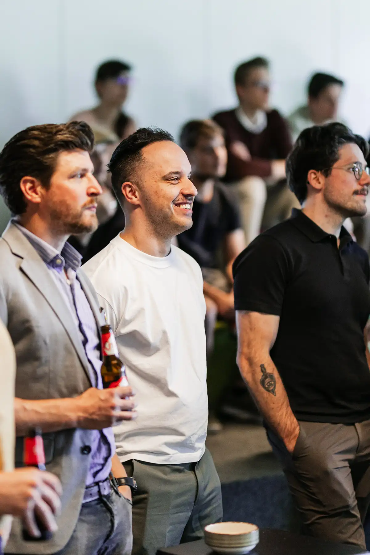 Three men stand together at a social gathering, smiling and chatting while holding drinks, with other people seated in the background.