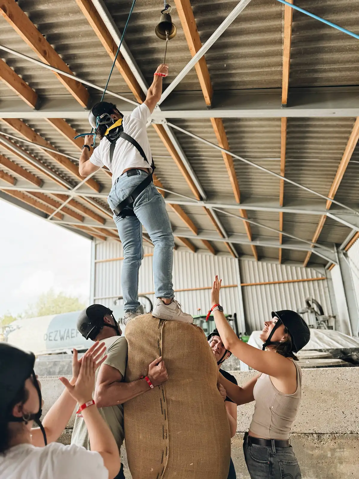 A person wearing a helmet and safety harness reaches up to ring a bell while standing on a large burlap-covered structure, surrounded by people assisting and spotting them.