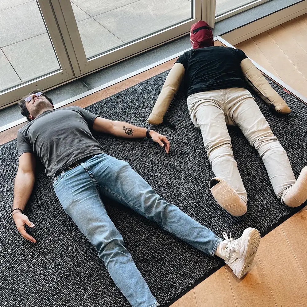 Two people lie on a carpeted floor near a glass entrance, appearing to participate in a playful or staged activity.