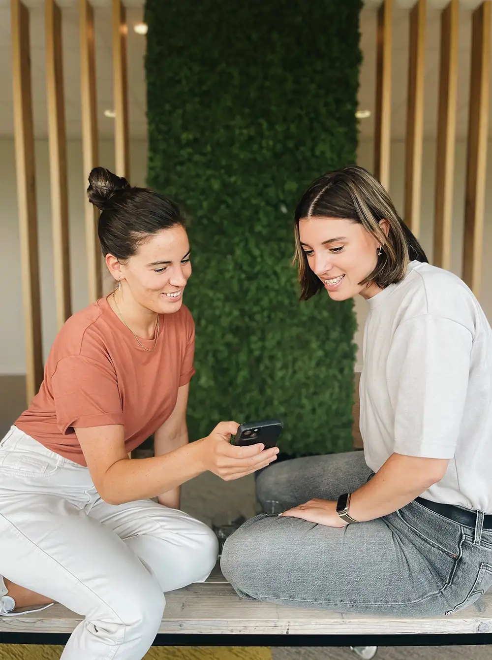 Two women sit together indoors, smiling as they look at a smartphone.
