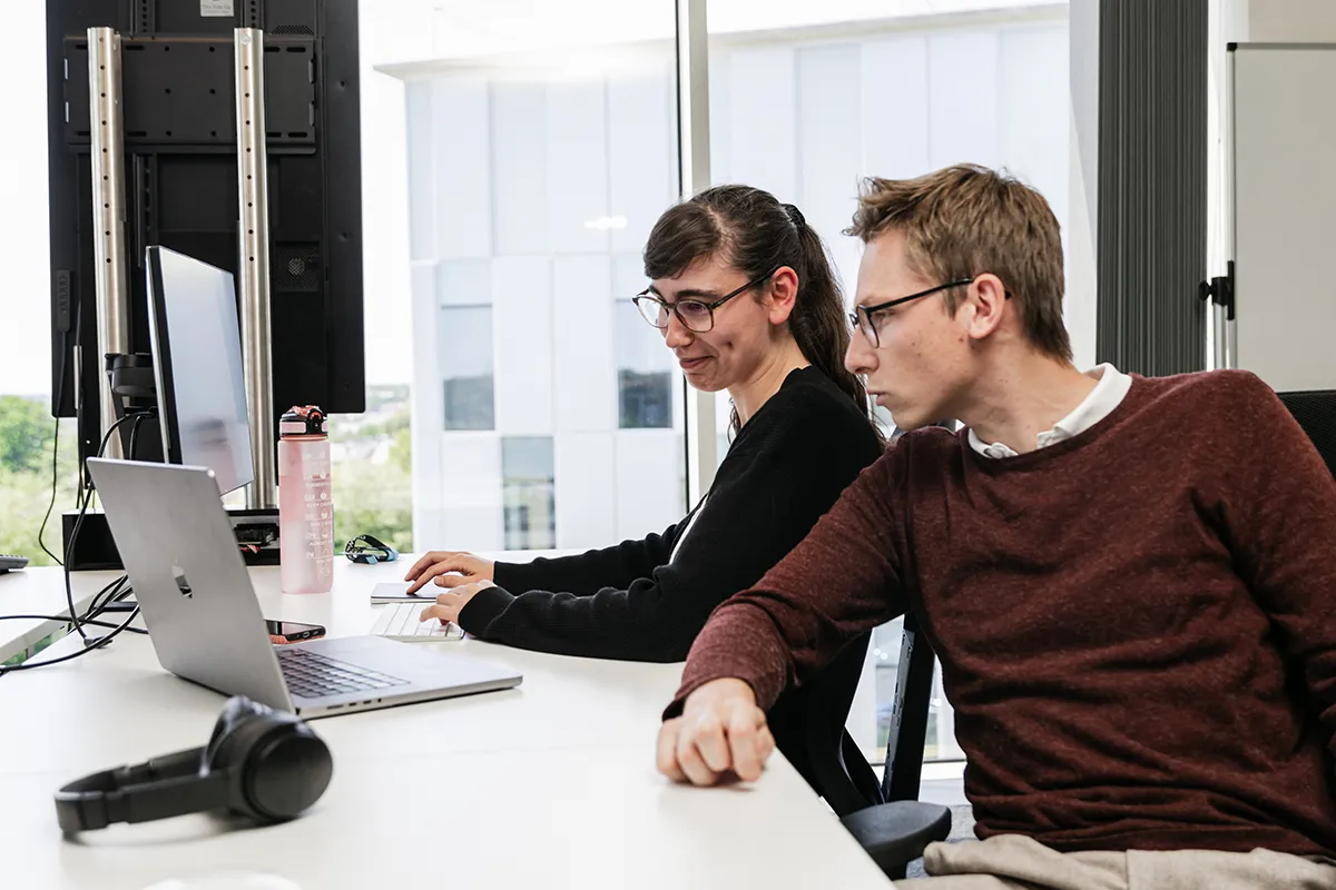 Two colleagues work together at a desk with laptops and monitors in a bright office.