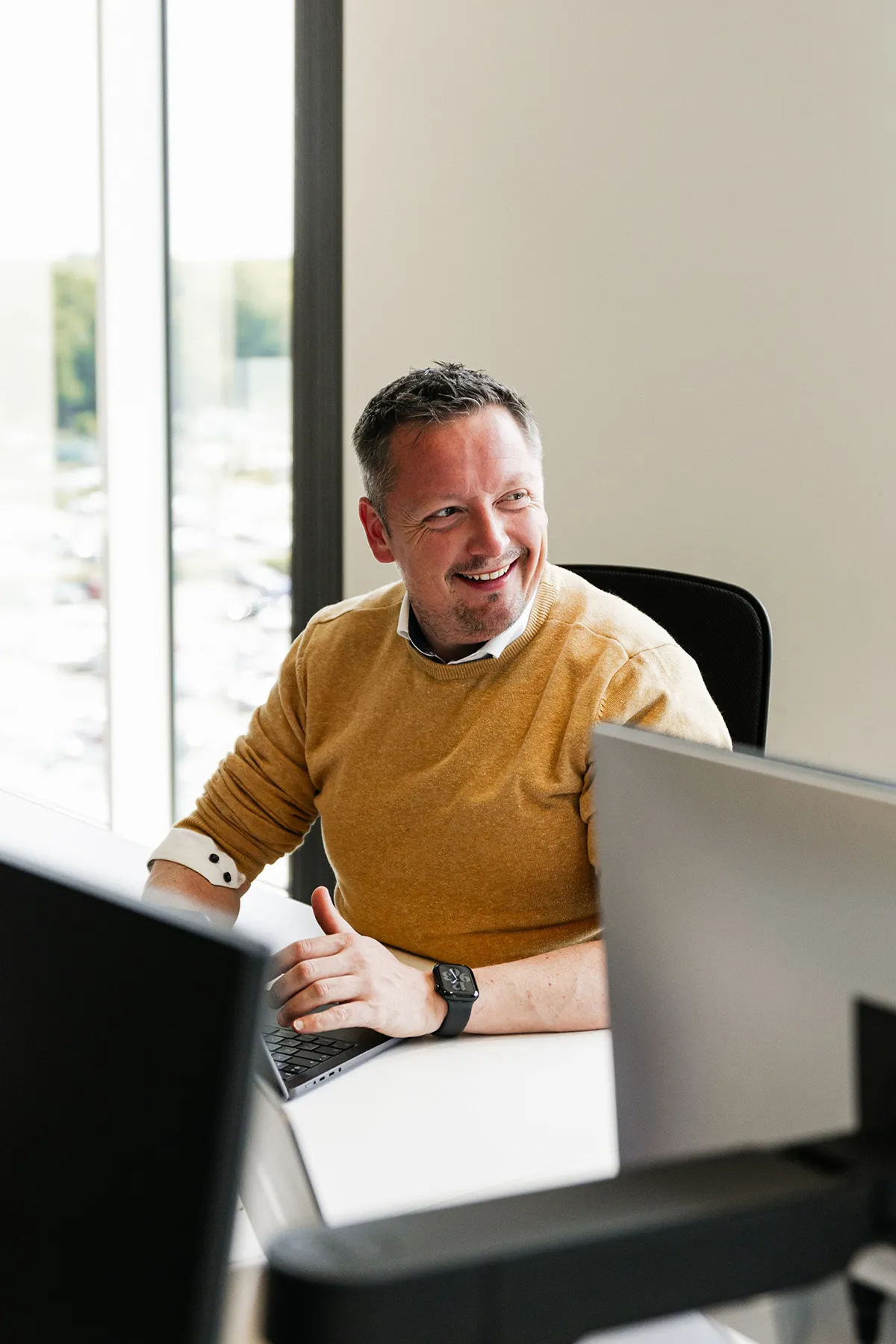A smiling man in a mustard sweater sits at his desk in front of computer monitors.