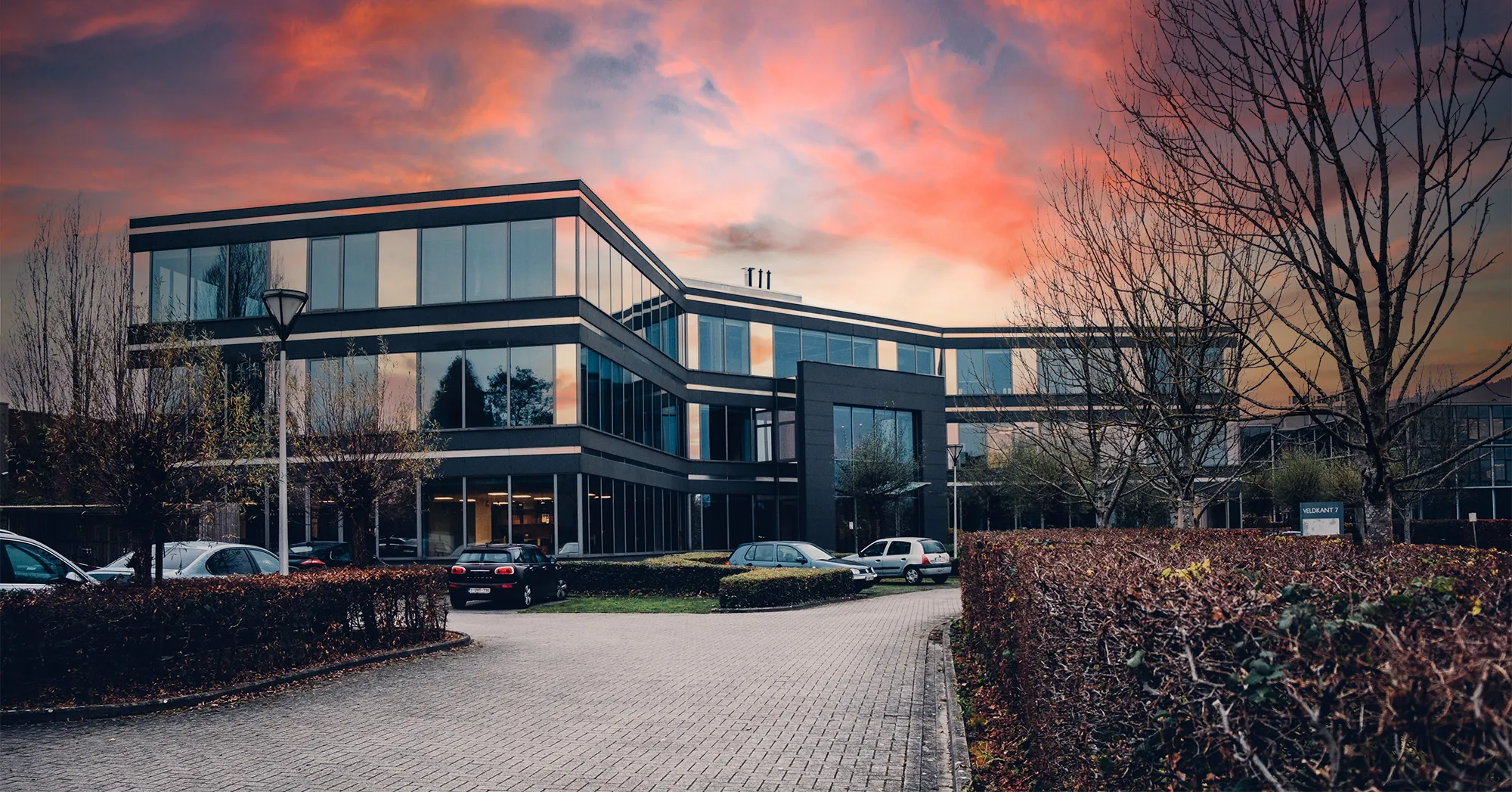 Modern office complex with large windows and dark façades, surrounded by trees and parked cars at sunset.