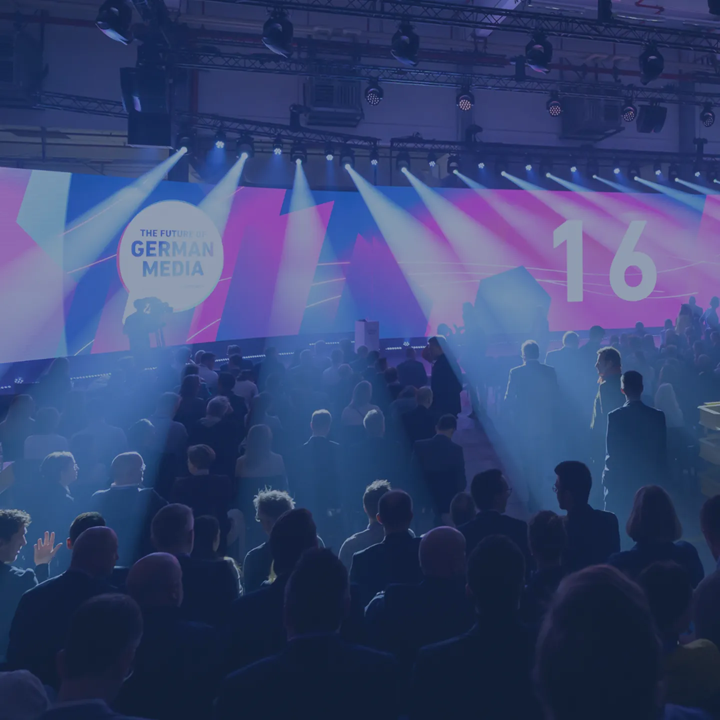Eine große Gruppe von Menschen sitzt und steht in einem abgedunkelten Konferenzsaal mit Bühnenbeleuchtung und einer großen Leinwand, auf der „The Future of German Media“ und die Zahl „16“ angezeigt werden.