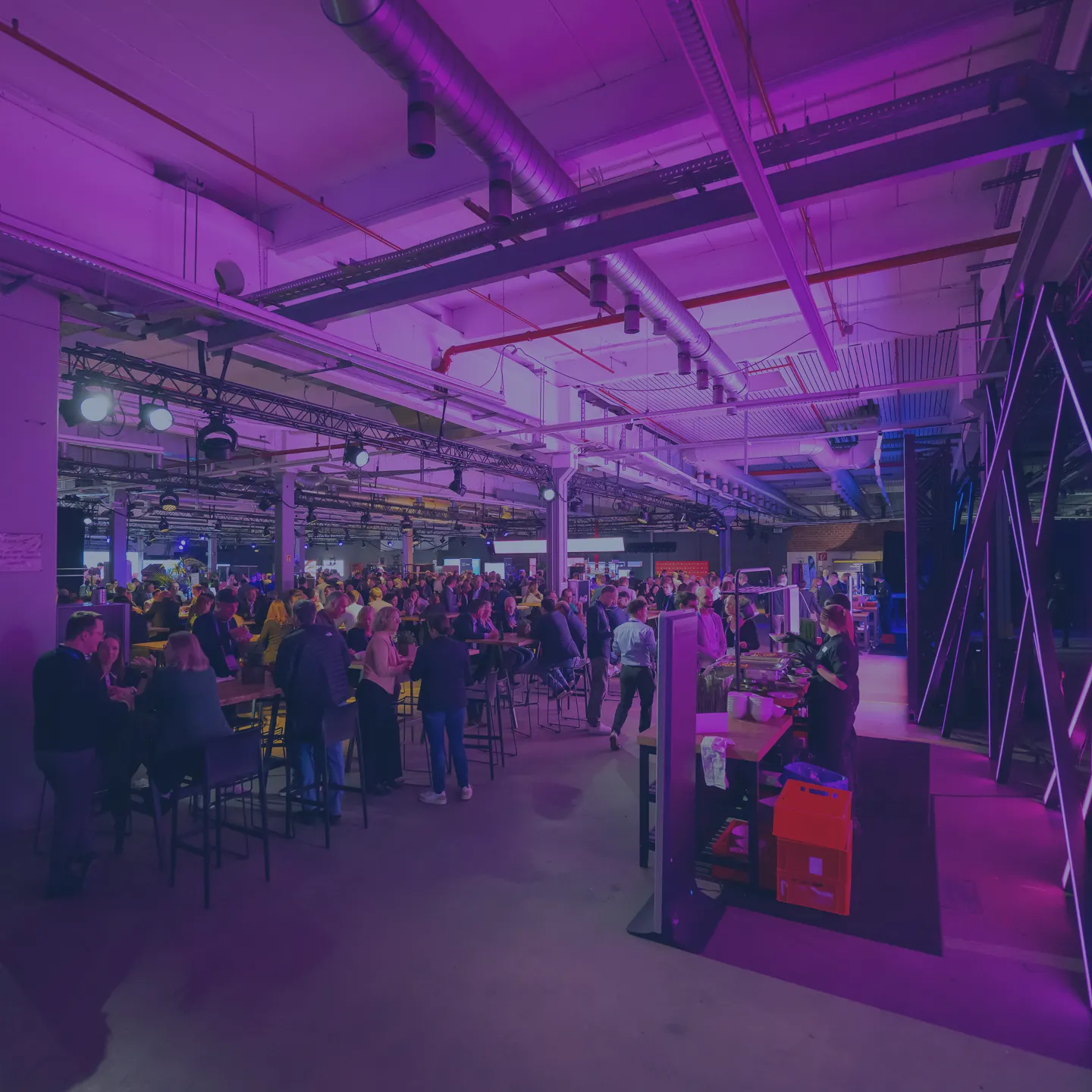 Menschen versammeln sich in einem großen, modernen Veranstaltungsraum mit Hochstühlen und Tischen, beleuchtet in lila Farbtönen, während ein Catering-Stand auf der rechten Seite Speisen anbietet.