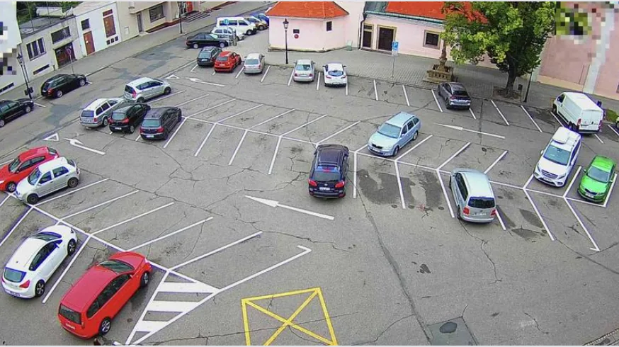Elevated camera view over a city surface car park with marked bays, parked cars and a few empty spaces