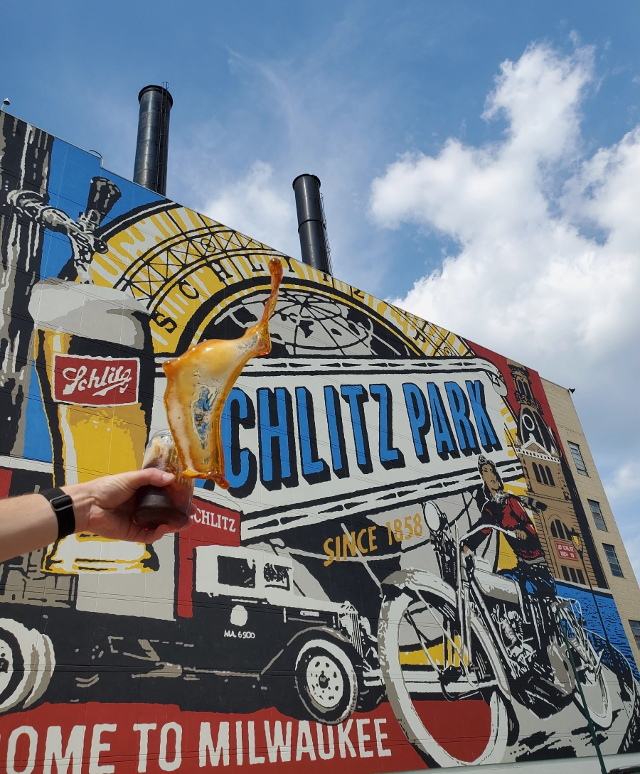 Photo of hand holding a beer in front of large Milwaukee Schlitz Park mural.