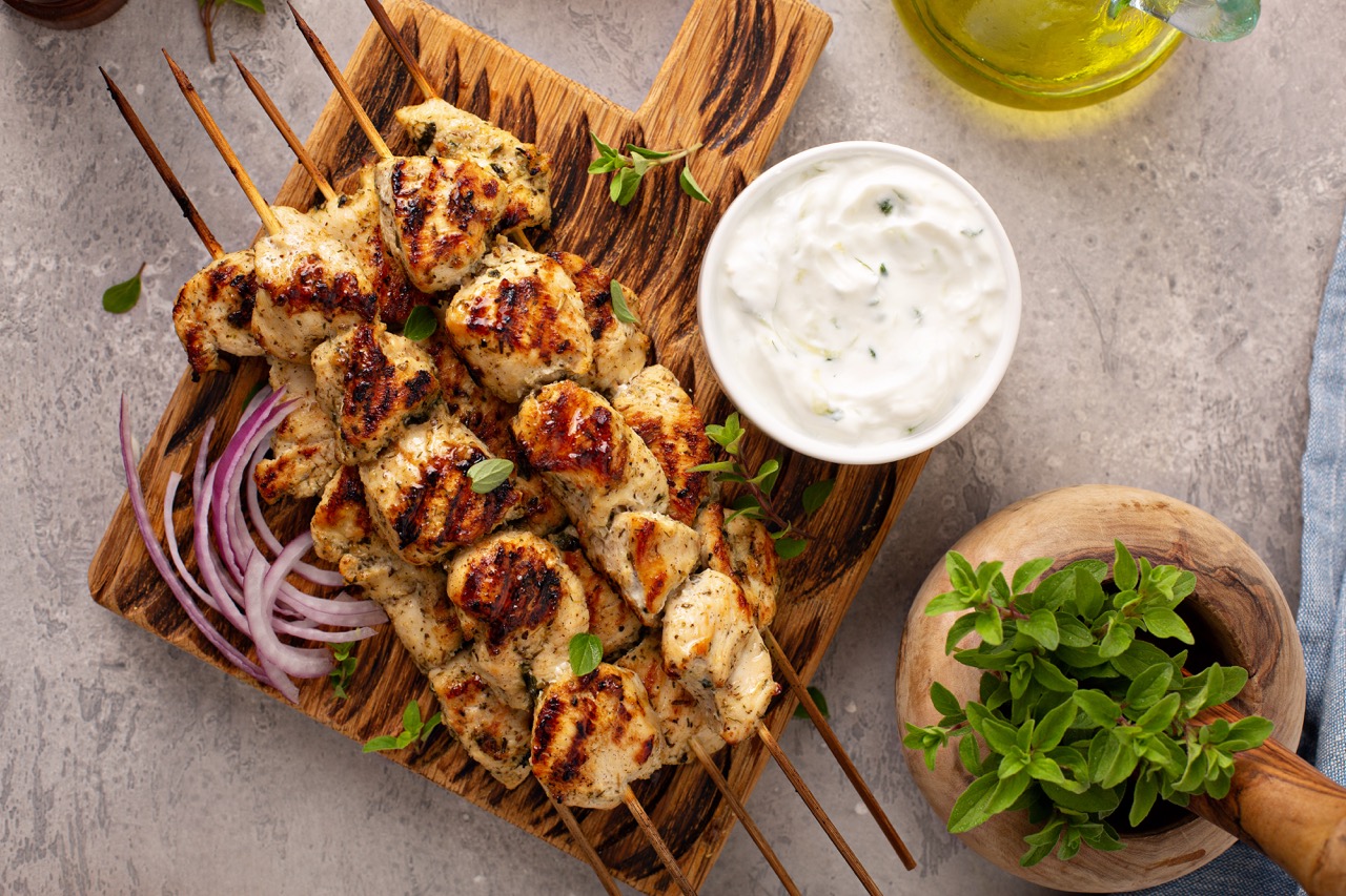 Grilled chicken skewers on a wooden cutting board garnished with fresh herbs, served with a bowl of creamy white sauce and sliced red onions, alongside a wooden bowl of green herbs and a bottle of olive oil on a gray surface.