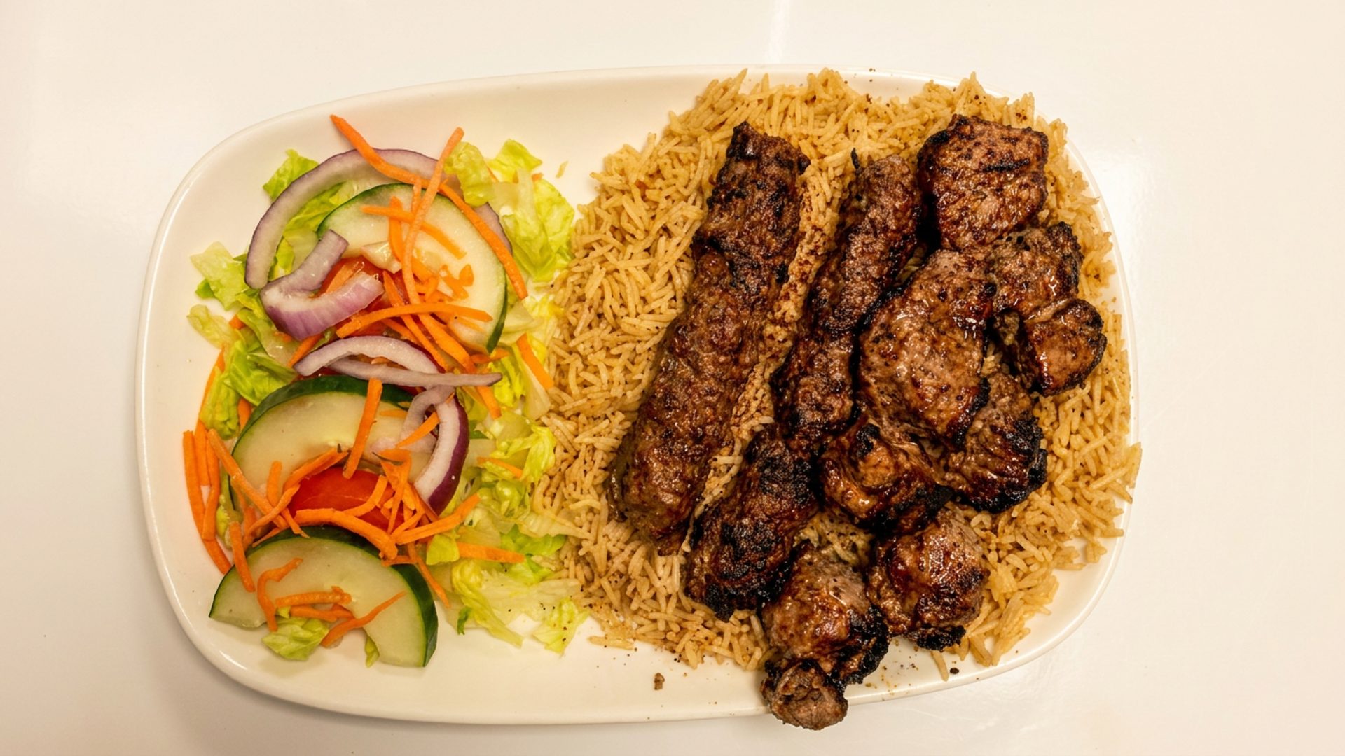 Plate with grilled meat skewers served over seasoned rice and a side salad of lettuce, cucumber slices, shredded carrots, and red onion rings.