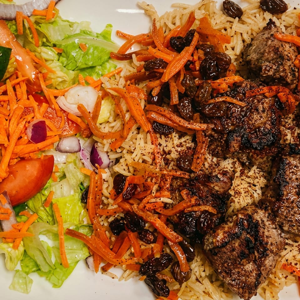 Plate with grilled meat cubes over seasoned rice topped with shredded carrots and raisins, alongside fresh salad with lettuce, tomato slices, shredded carrots, and chopped red onions.