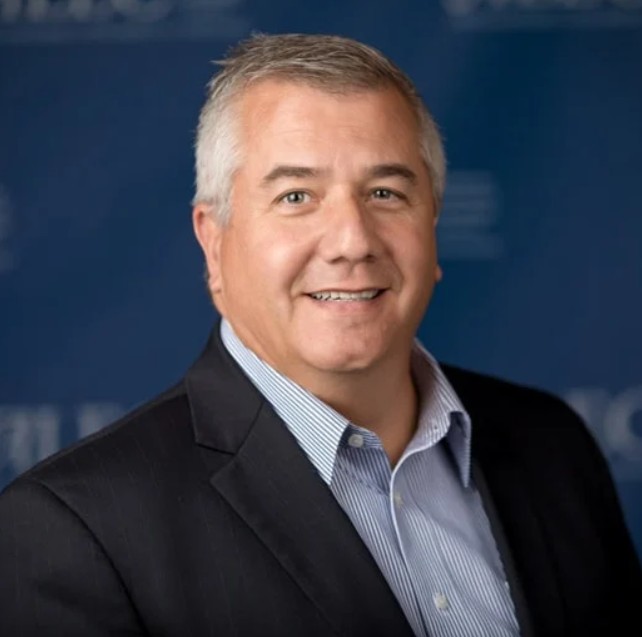 Smiling middle-aged man with short gray hair wearing a dark blazer and light blue striped shirt against a blurred blue background.