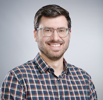 Man with short dark hair, beard, and glasses wearing a blue and red checkered shirt smiling against a plain light gray background.