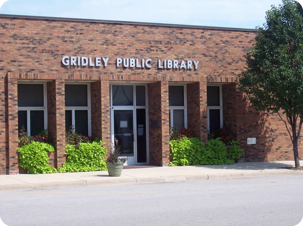 Front view of Gridley Public Library, a one-story brick building with four vertical windows flanking a central glass door, green plants in front, and a tree on the right side.
