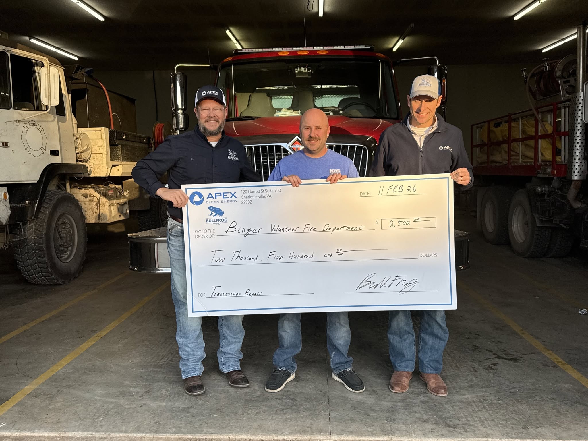 Three men standing in a fire station holding a large check for $2,500 made out to Binger Volunteer Fire Department for transmission repair, with a red fire truck and equipment trucks in the background.