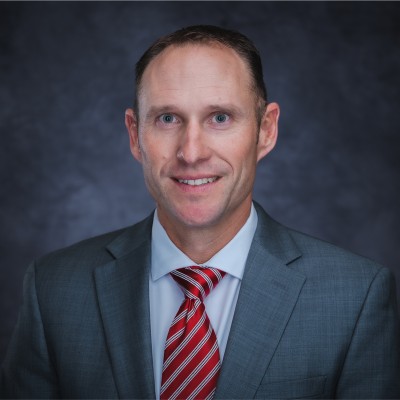 Portrait of a man with short brown hair and blue eyes wearing a gray suit, white shirt, and red striped tie, smiling against a dark gray blurred background.