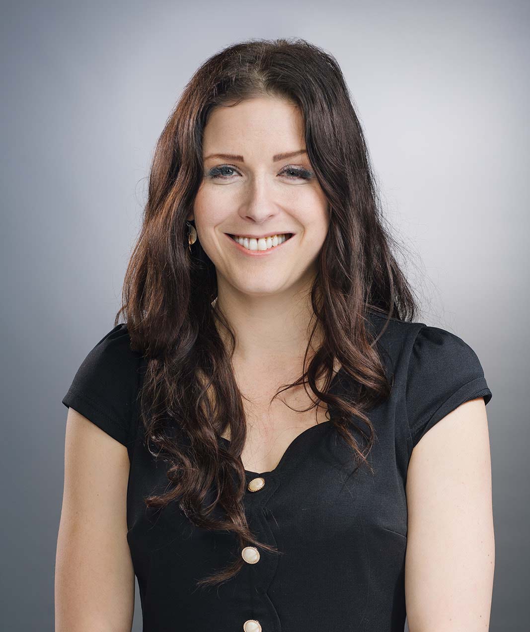 Smiling woman with long wavy brown hair wearing a black buttoned top against a gray gradient background.