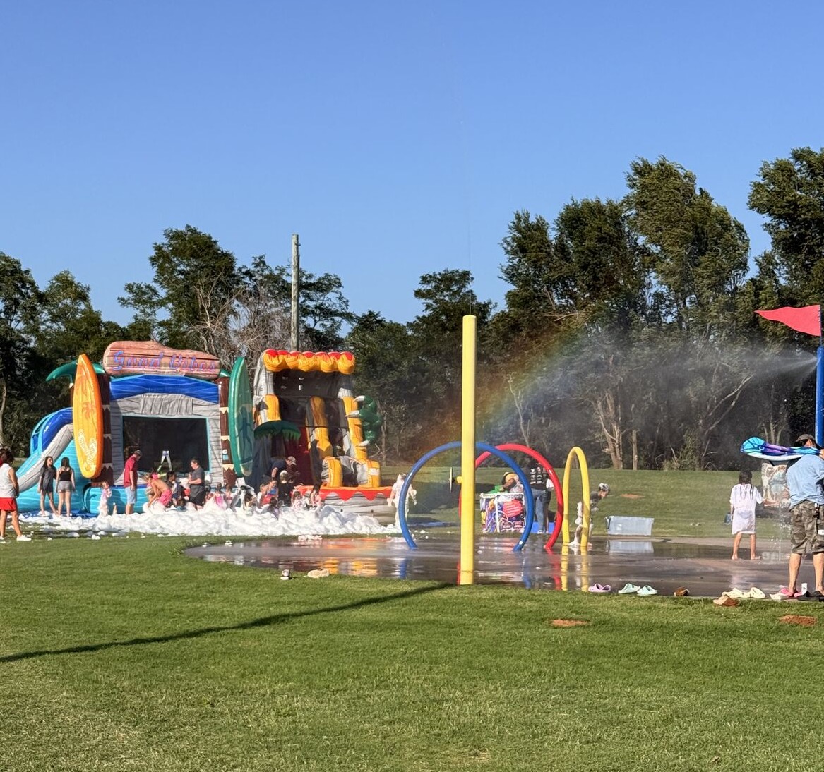 Outdoor park scene on a sunny day with children and adults playing near inflatable water slides surrounded by foam and a splash pad with colored hoops spraying water, and a faint rainbow visible in the spray.