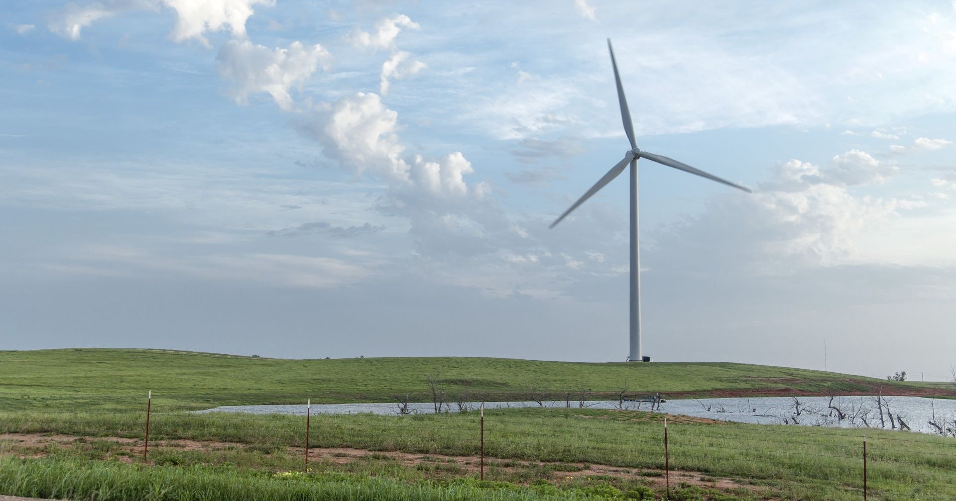 Single wind turbine with spinning blades on a grassy hill near a small pond under a partly cloudy sky.