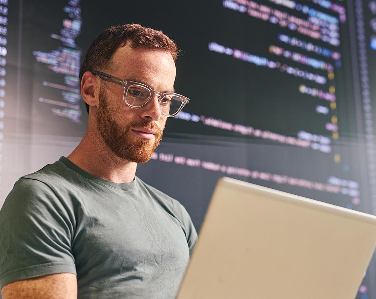 A man works on a laptop in front of screens displaying code.