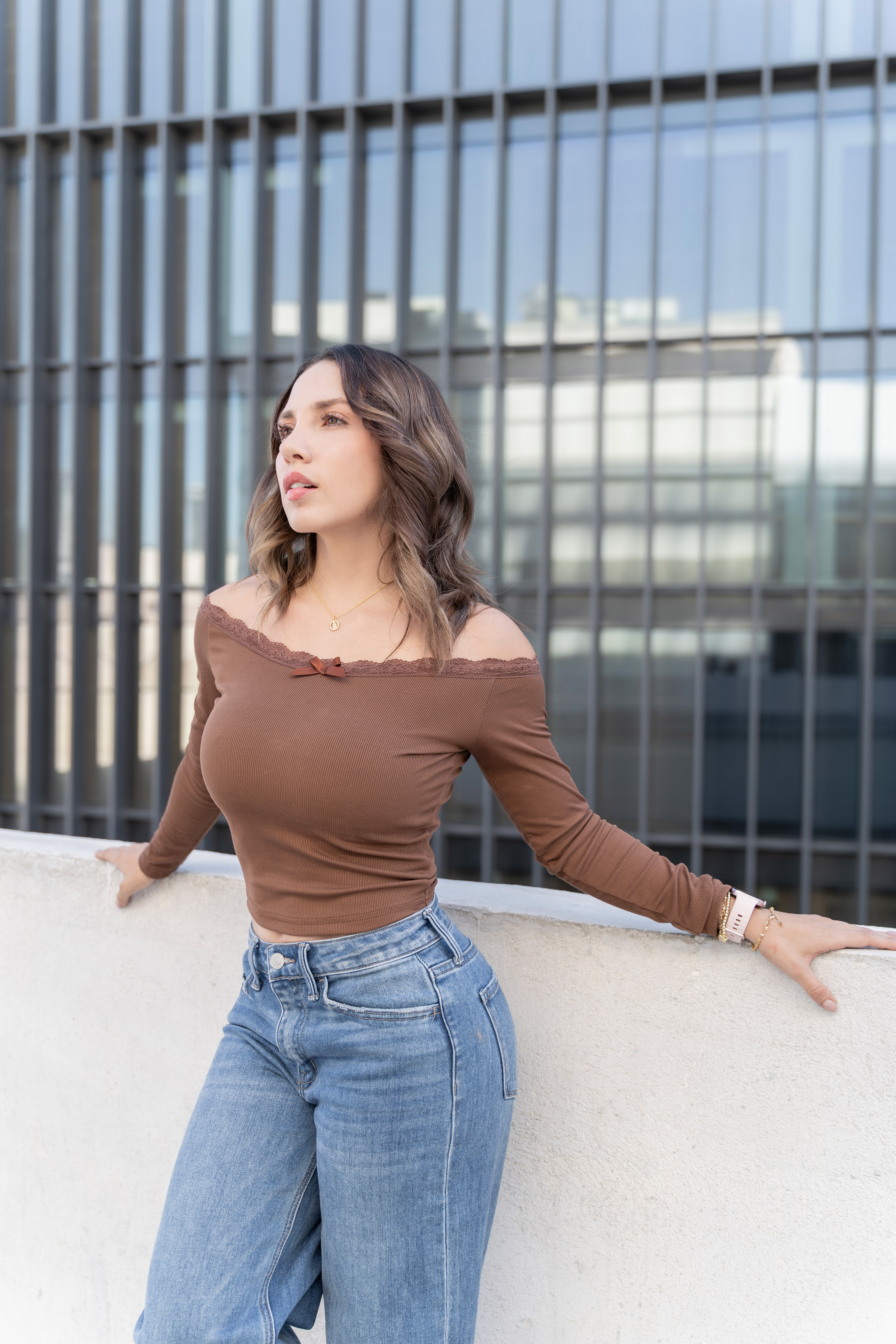 Mujer con blusa café recargada en un muro frente a un edificio de cristal