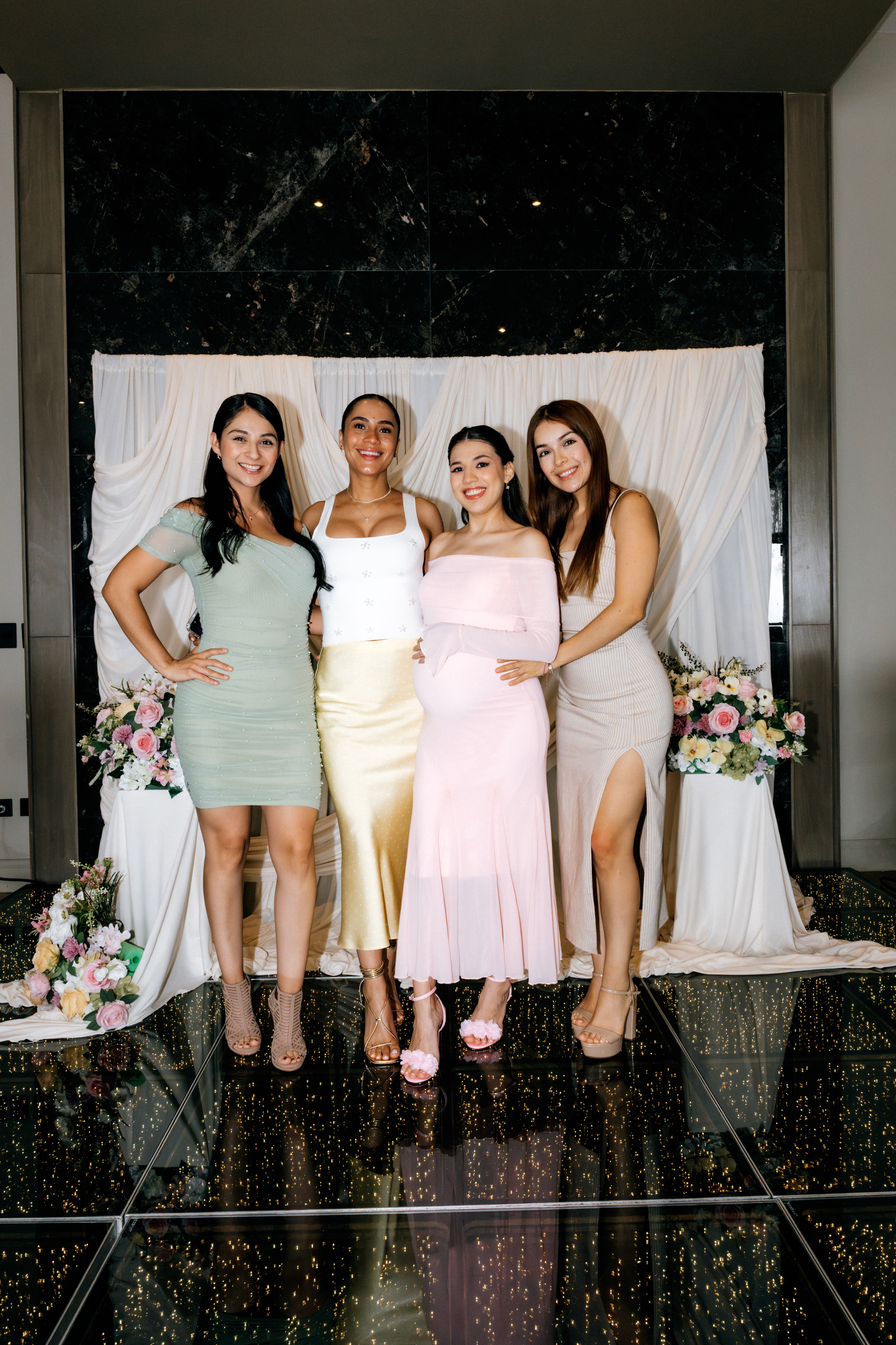 Cuatro mujeres posando frente a un fondo de tela blanca con flores en una celebración