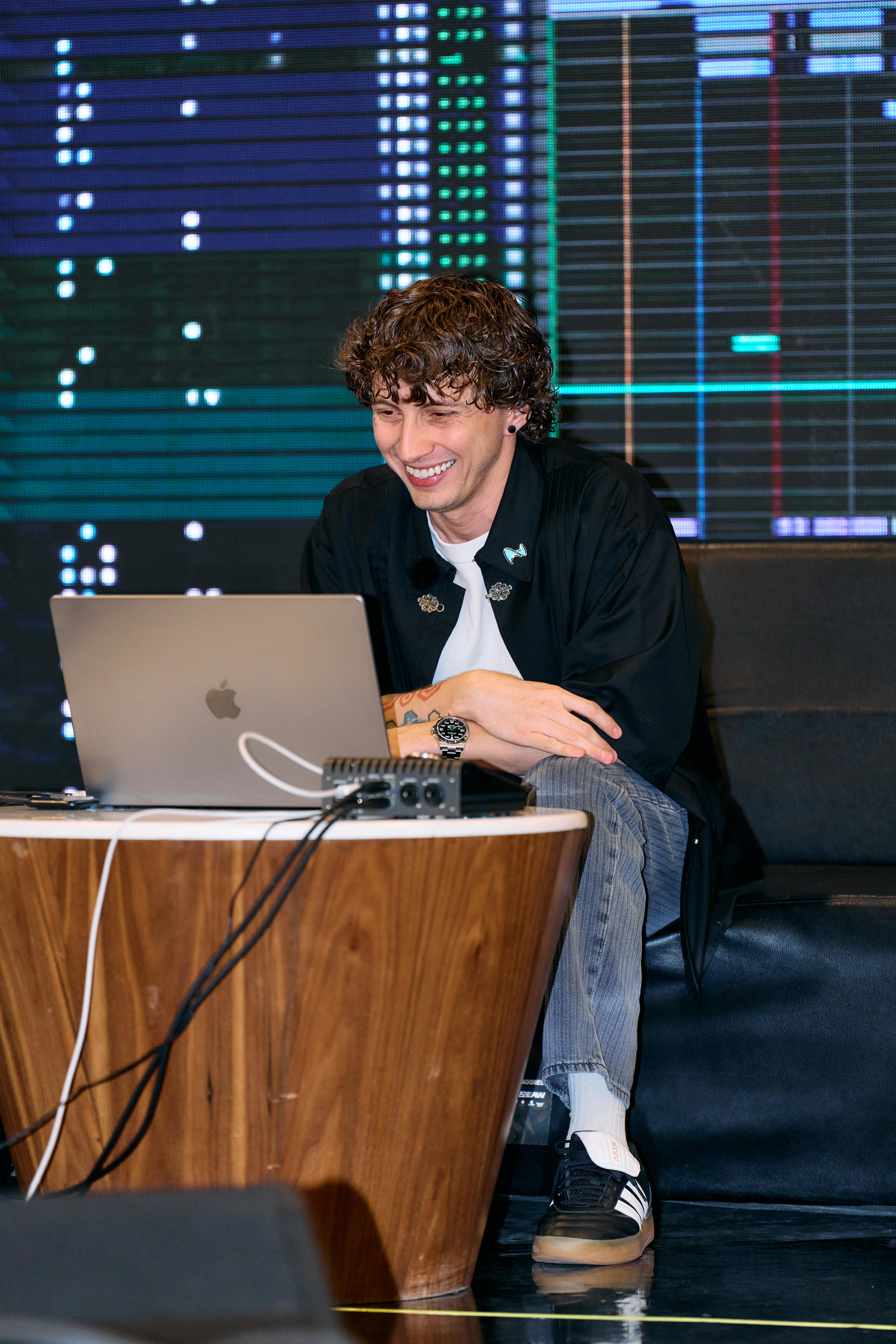 Joven sonriendo frente a una laptop durante un panel, con una pantalla de luces detrás