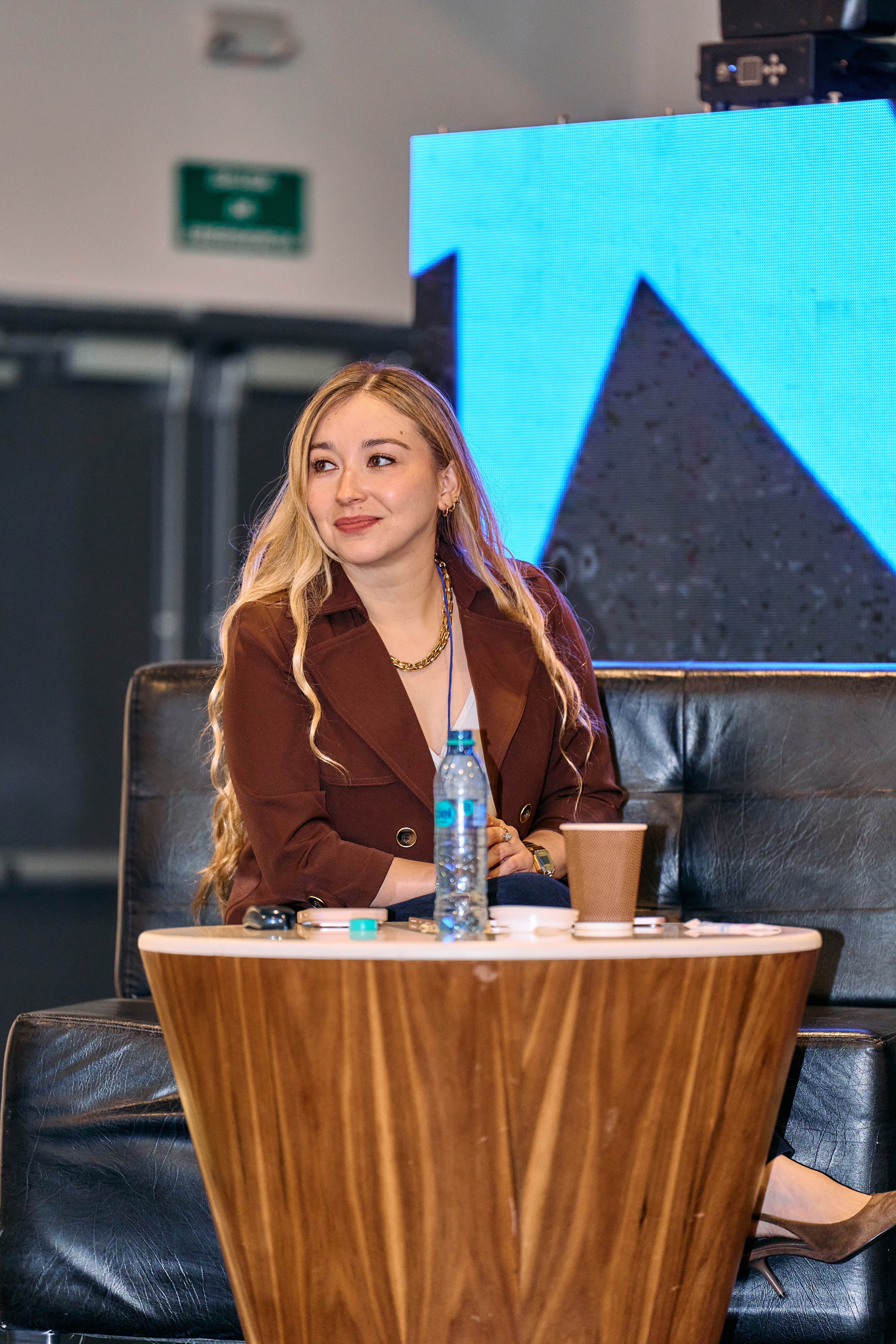 Mujer sonriendo sentada en un sillón durante un panel, con un vaso de agua en la mesa