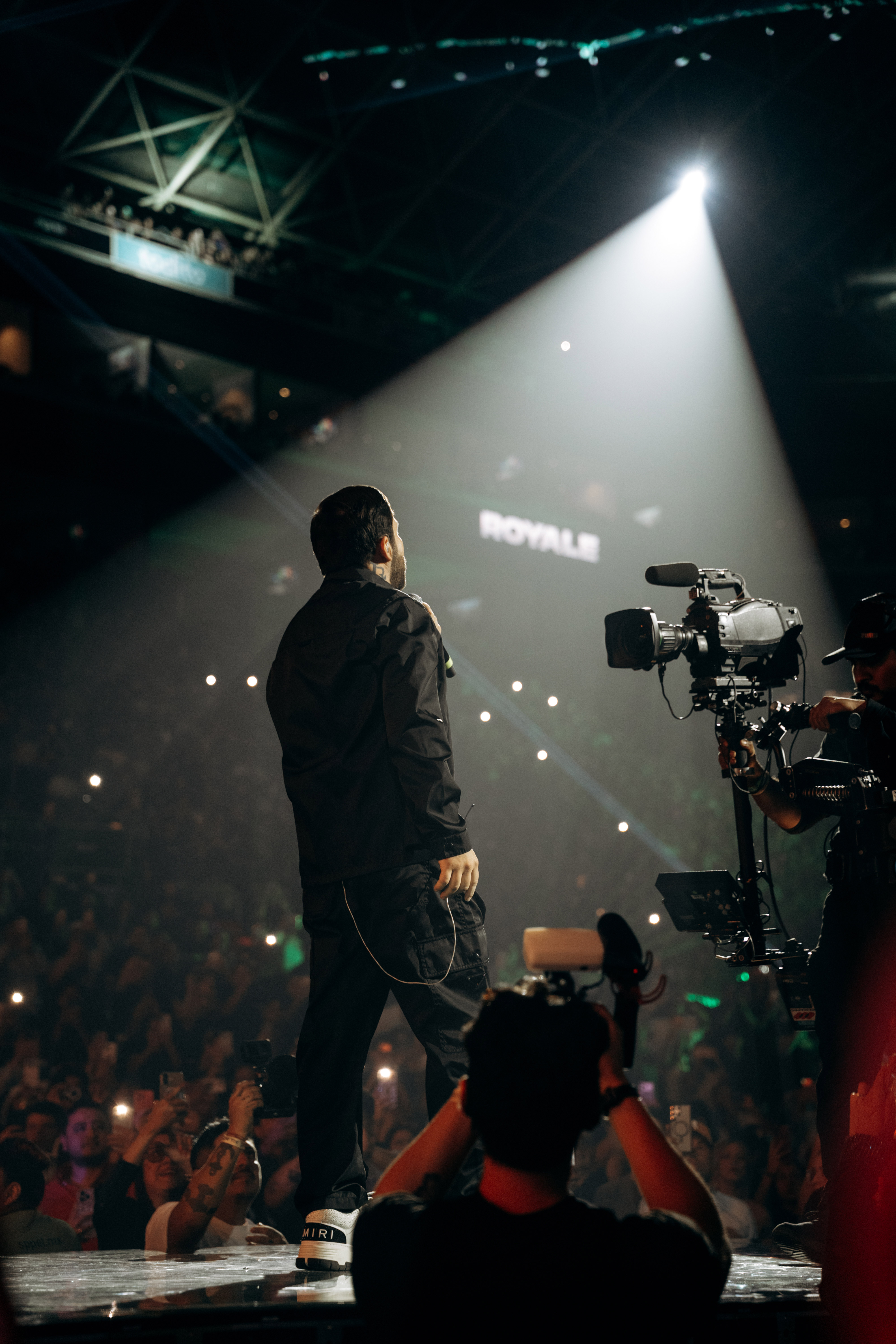 Persona de espaldas en el escenario de Ring Royale frente a miles de asistentes, junto a cámaras de video
