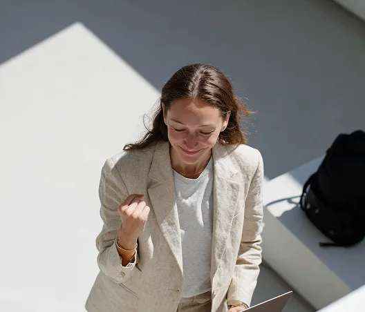 Smiling woman in beige suit looks down while clenching fist in a gesture of success.