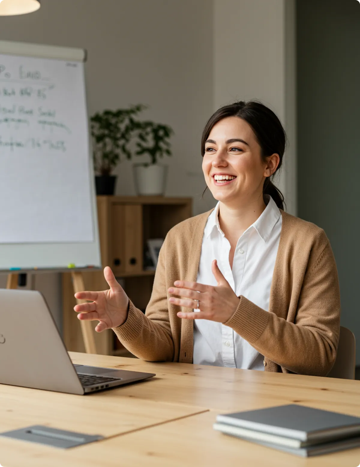 Woman smiling and gesturing while sitting at a wooden table with a laptop and notebooks.