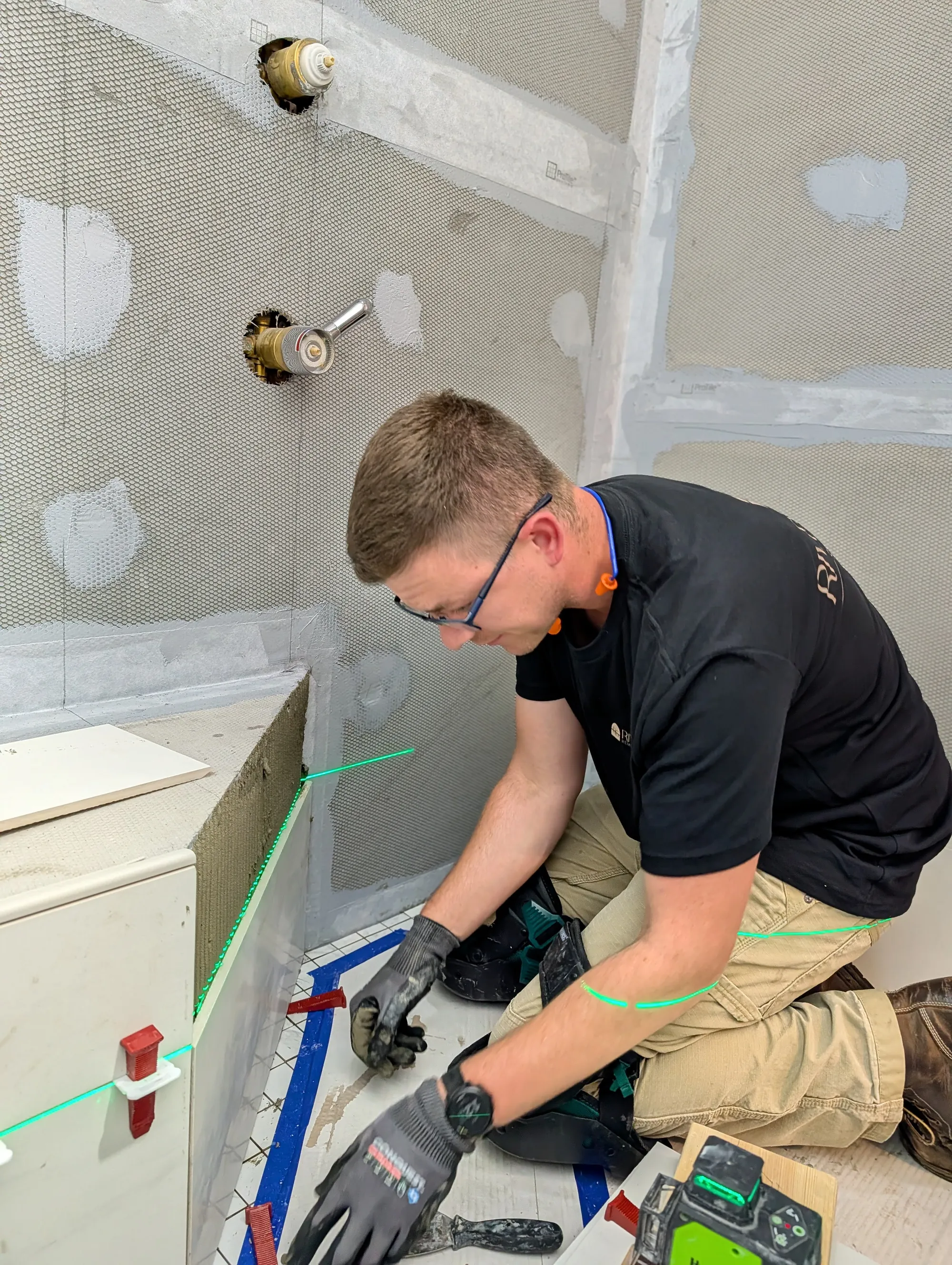 Austin Lapp in safety glasses and work gloves aligning shower wall tile to a green laser line with tile spacers