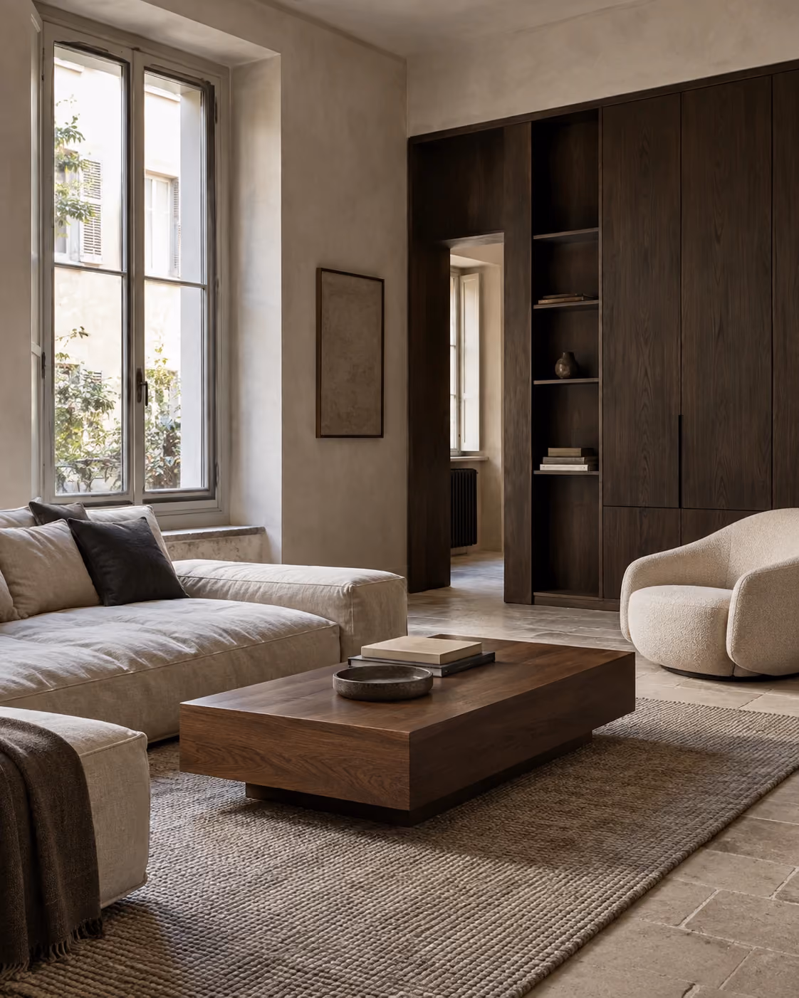 Minimalist living room with neutral tones featuring a large beige sectional sofa with cushions, a low rectangular wooden coffee table on a textured rug, a rounded cream armchair, large window letting in natural light, and dark wood built-in shelves and cabinets.